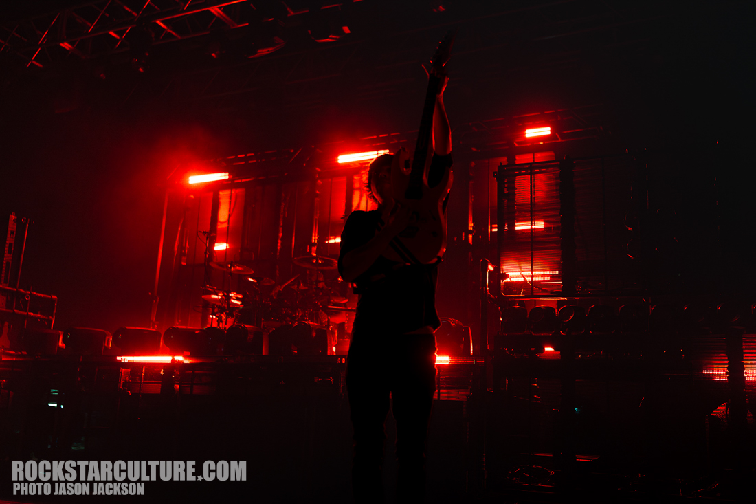 A silhouetted musician holds a guitar high in the air on stage, with dramatic red lighting and equipment visible in the background.