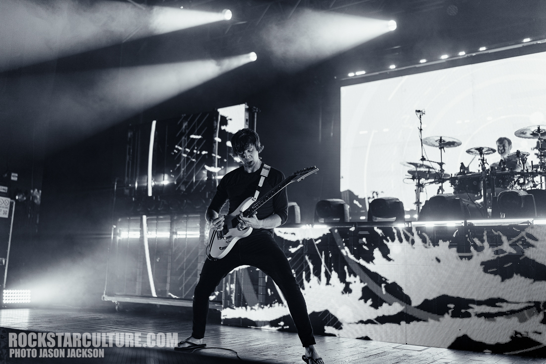 A musician performing on stage with an electric guitar, surrounded by dramatic lighting effects and a backdrop of screens displaying abstract patterns.