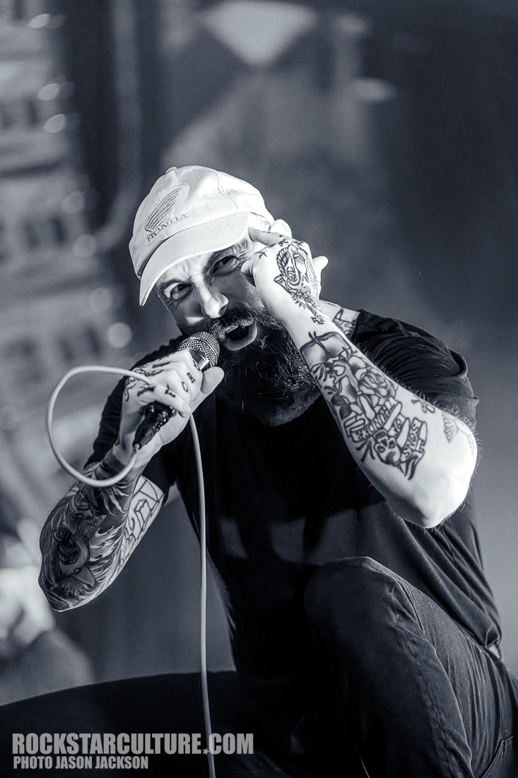 A tattooed male vocalist passionately singing into a microphone while crouching on stage, wearing a white cap and a black t-shirt.
