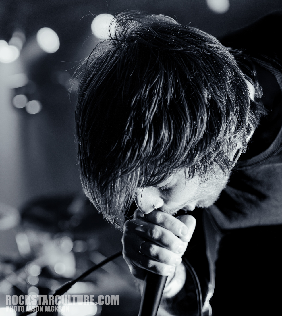 A close-up shot of a male singer passionately performing into a microphone, with his eyes closed and hair falling over his face, set against a blurred background of stage lights.