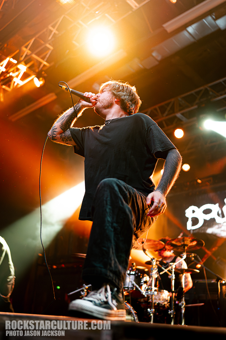 A vocalist passionately performing on stage with a microphone during a live concert, illuminated by stage lights.