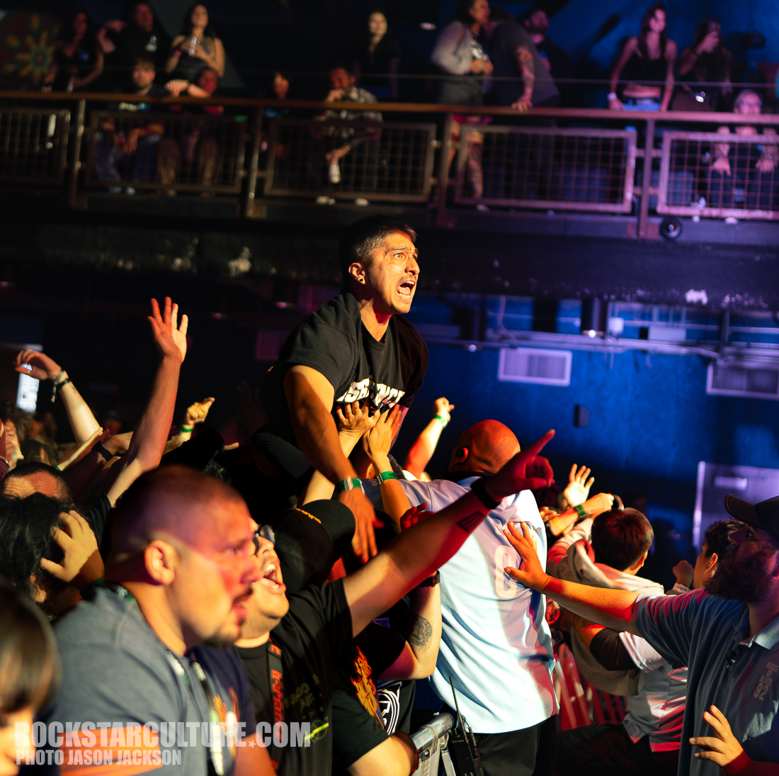A crowd of enthusiastic concertgoers with hands raised, some catching a person crowd surfing above them during a live performance.