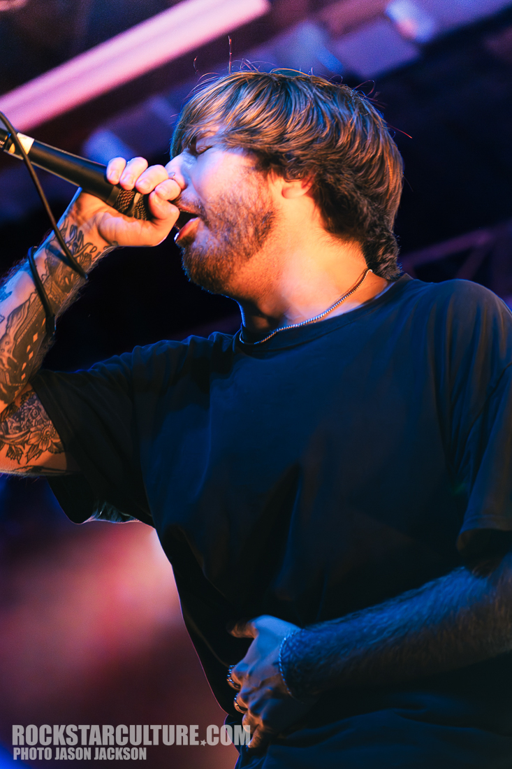 A singer passionately performing on stage with a microphone, wearing a black t-shirt and showcasing tattooed arms.