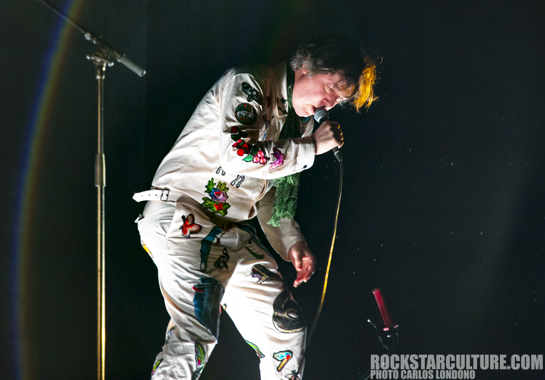 A performer singing into a microphone while wearing a white outfit adorned with colorful floral and animal embroidery.