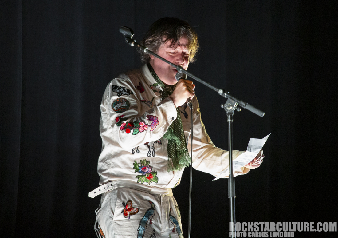 A performer stands at a microphone on stage, wearing a brightly embroidered outfit with floral and animal designs, holding a piece of paper and speaking to the audience.