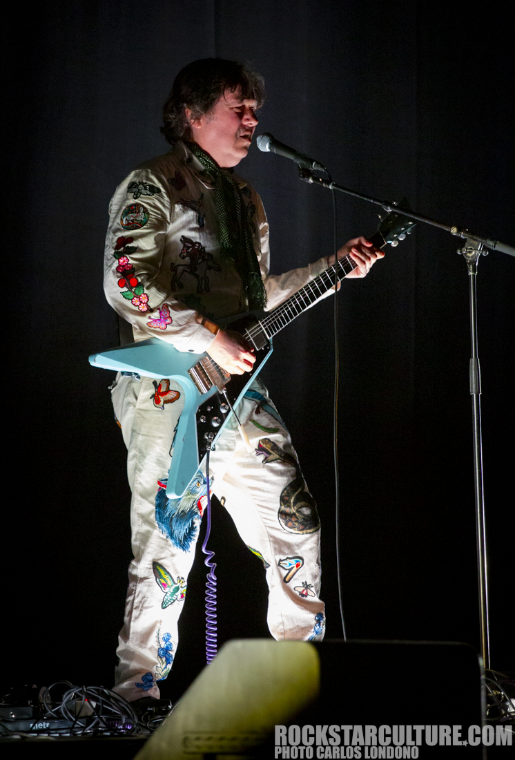A musician performing on stage, wearing an embroidered jumpsuit and playing a light blue guitar.