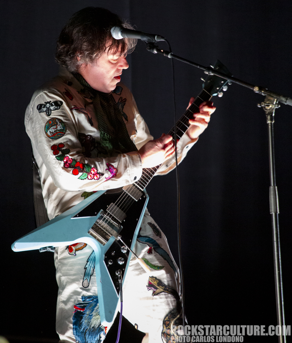 A musician with shoulder-length hair performs on stage, wearing a white shirt adorned with colorful embroidery. He plays a blue electric guitar while leaning forward, showing focus and passion during the performance.