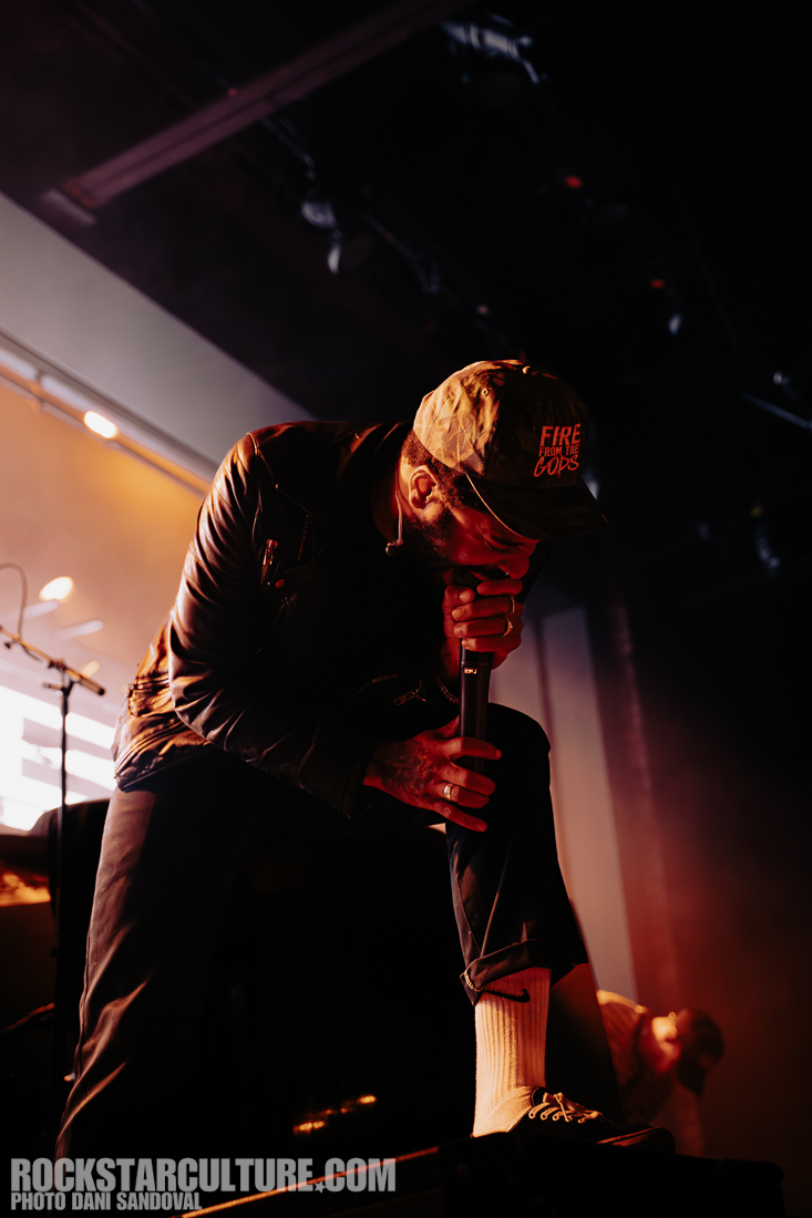 A musician performing on stage, holding a microphone and wearing a cap, with dramatic lighting and a leather jacket.