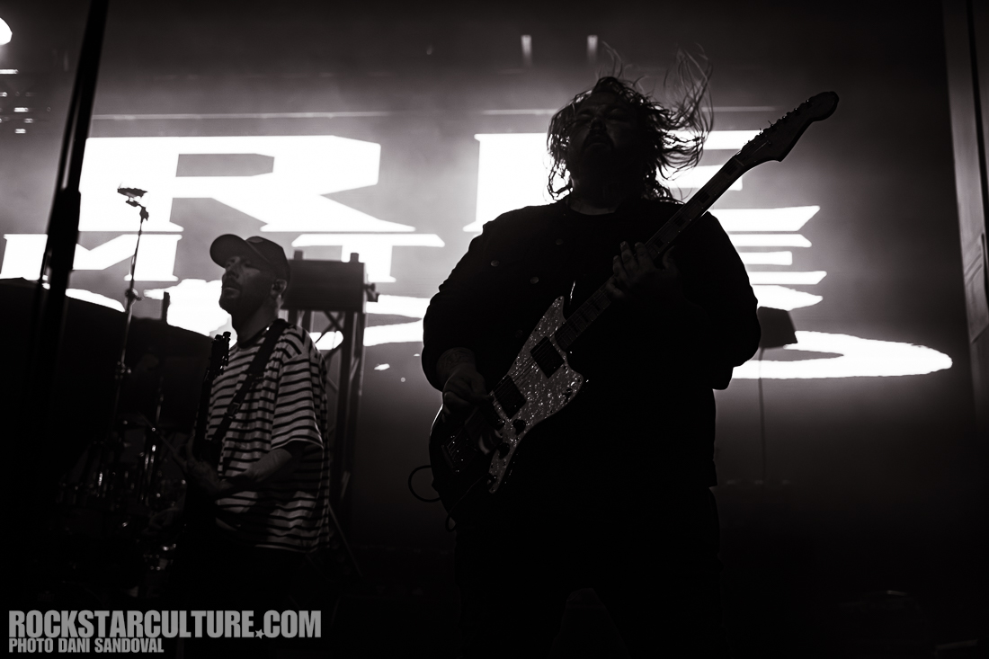 Silhouetted musicians performing on stage with dynamic lighting, one is playing guitar and another is visible on drums, captured in black and white.