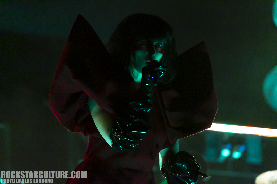 A performer sings into a microphone, dressed in a striking red outfit with large, dramatic sleeves and wearing black gloves, set against a dark, moody backdrop with green lighting.