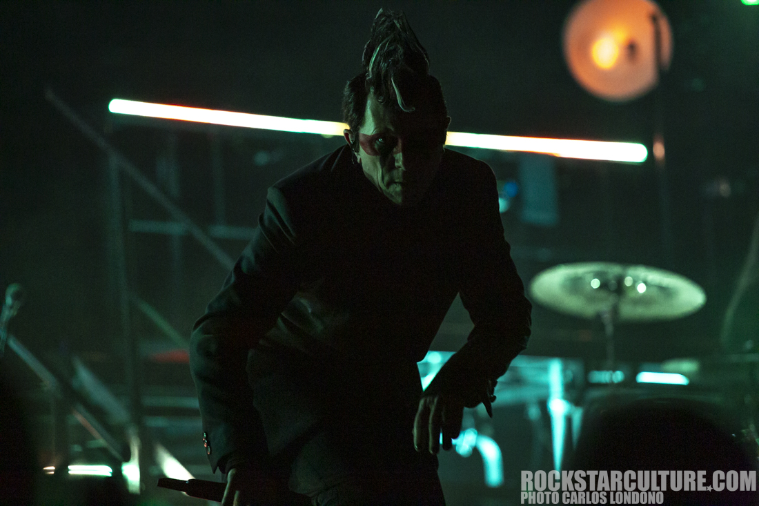 A performer on stage with dramatic lighting, wearing a black suit and a unique hairstyle, captured in a moody atmosphere.