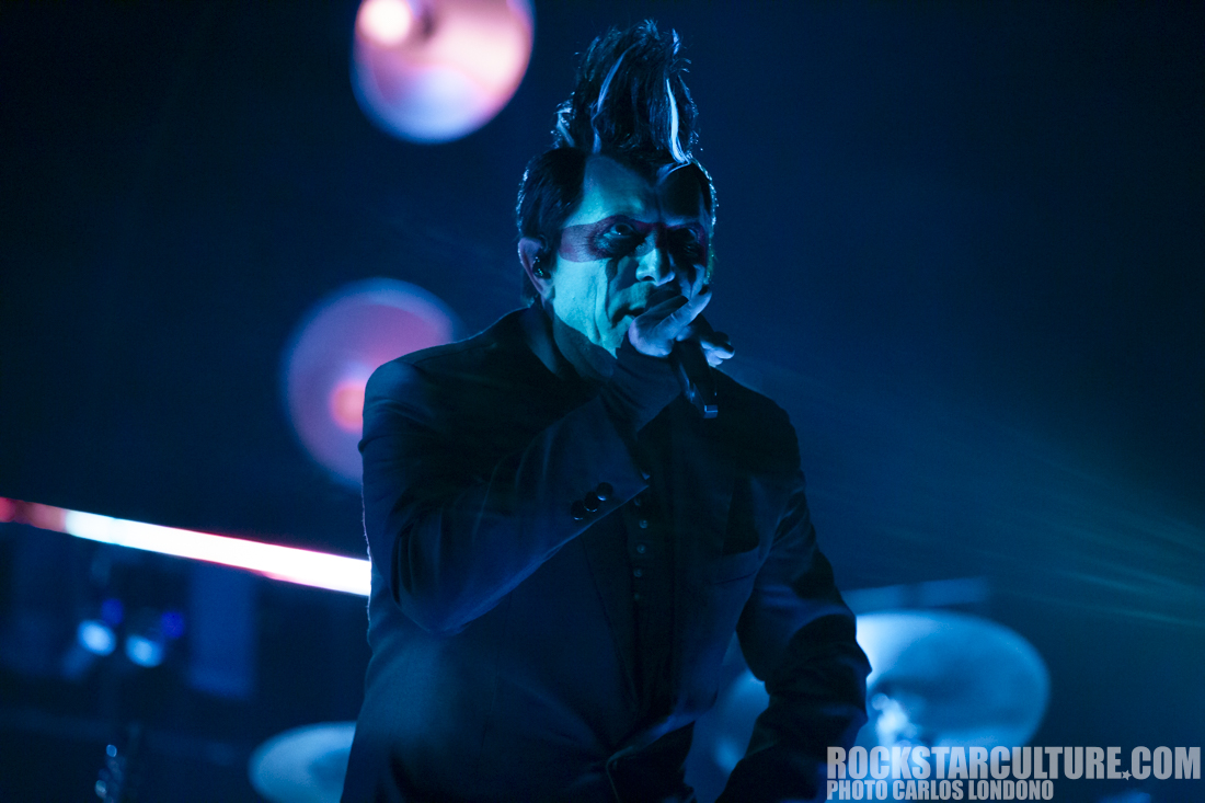 A singer with a striking mohawk hairstyle performs on stage, wearing a black jacket and face paint, set against a dimly lit background with colorful lights.