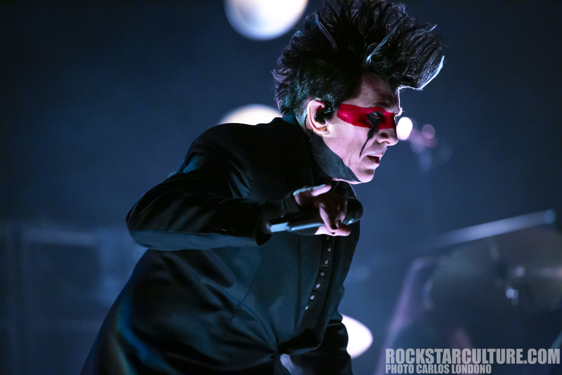 A performer with dramatic makeup and spiked hair, singing passionately into a microphone on stage, with blue lighting creating a vibrant atmosphere.