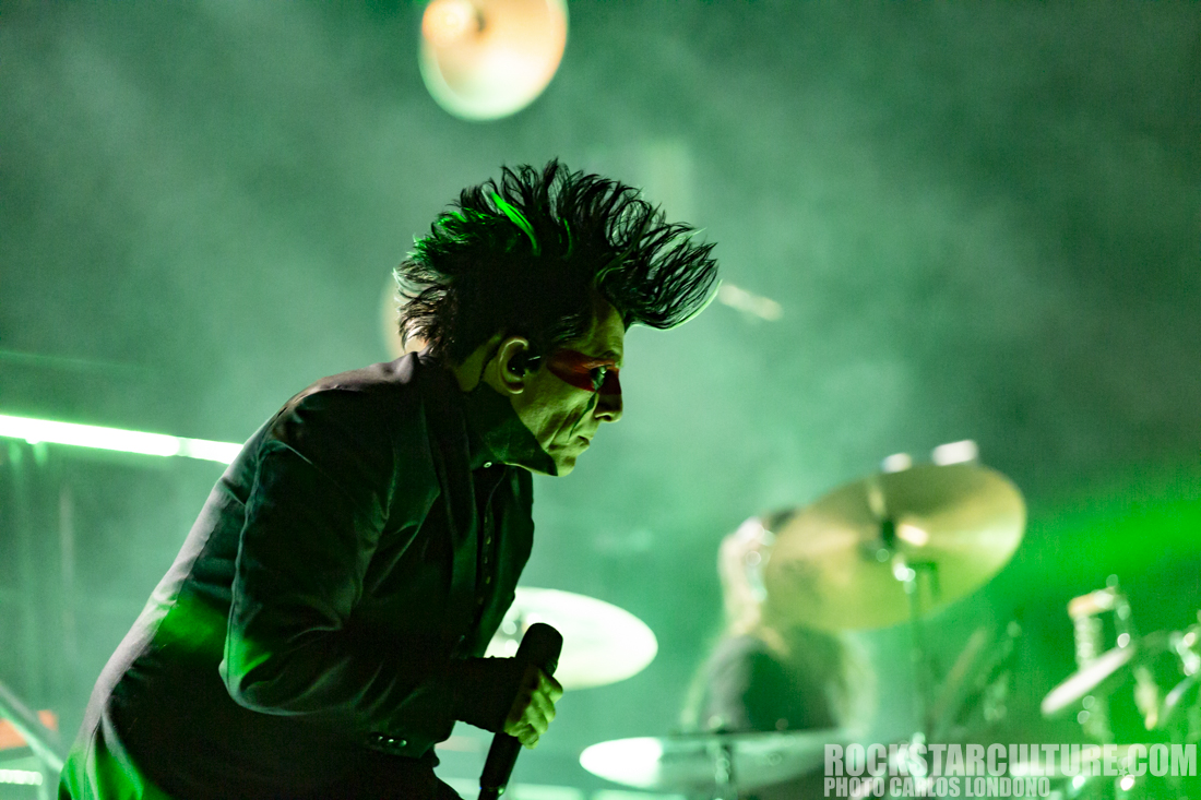A performer with spiky hair and sunglasses sings passionately on stage amidst green lighting, with a drummer partially visible in the background.
