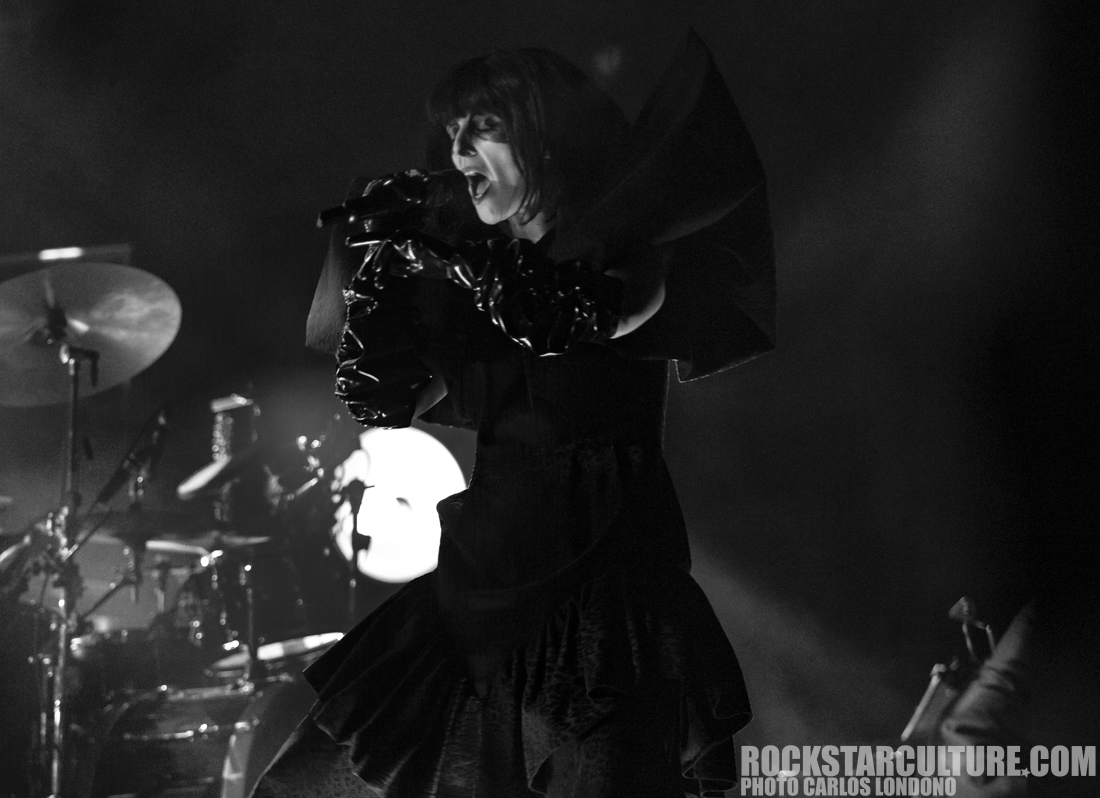 A singer performing on stage, dressed in a dramatic black outfit with oversized sleeves, in a dimly lit venue with a drum set visible in the background. The image is in black and white.