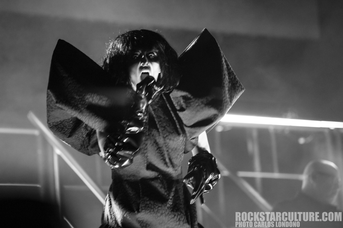 A performer in a dramatic outfit with large sleeves and dark, wild hair, passionately singing on stage, set against a moody black and white backdrop.