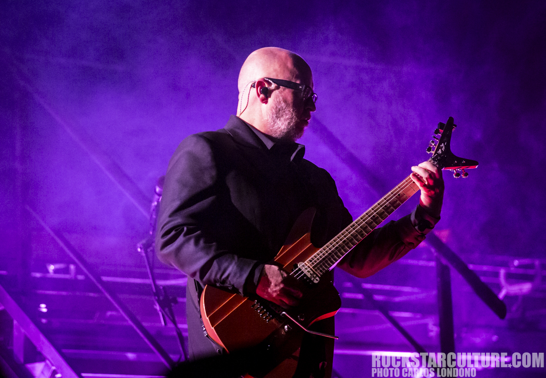 A musician performing on stage with an electric guitar under purple lighting.