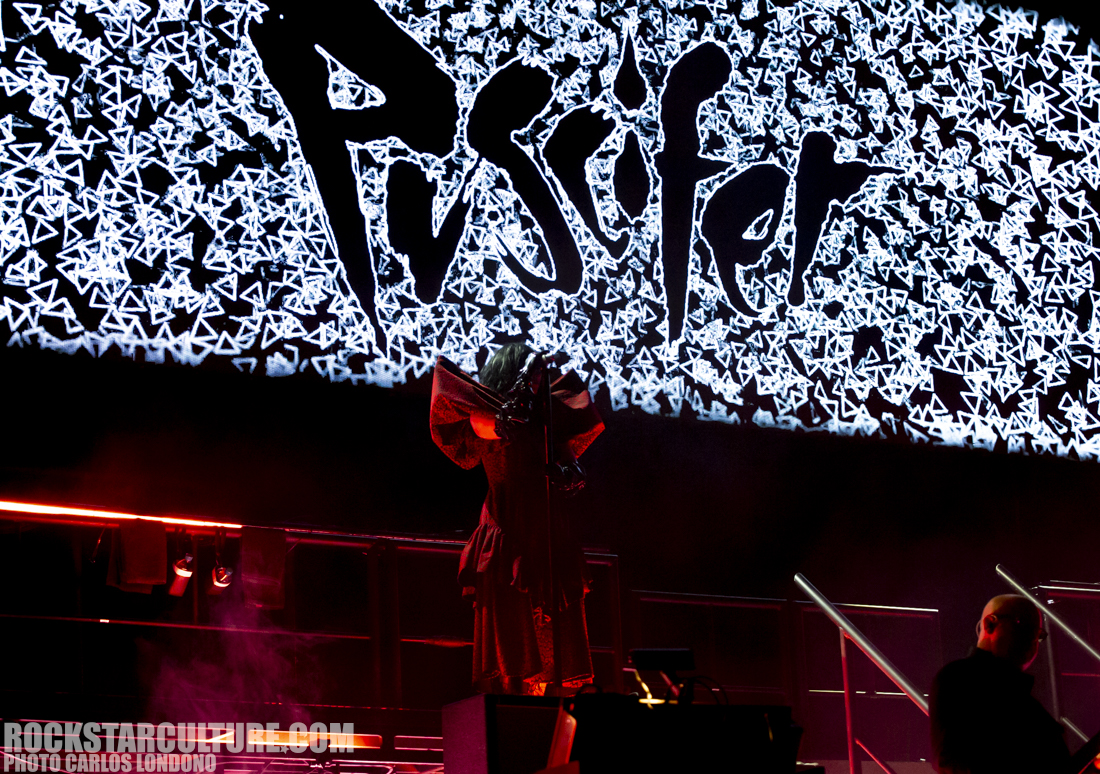 A performer on stage in front of a large illuminated backdrop displaying the name 'Puscifer' in a striking design, with vibrant lighting and smoke effects.