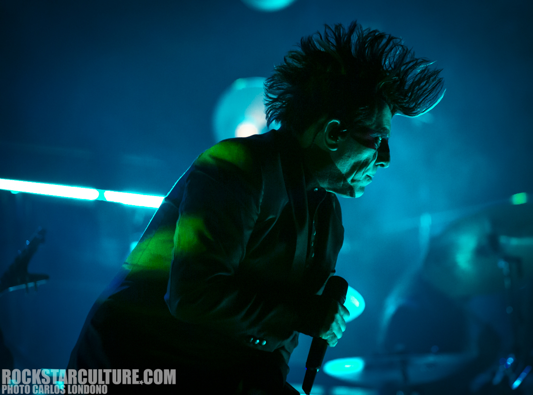 A musician performing on stage, captured in a dramatic silhouette with spiky hair, holding a microphone under blue and green stage lights.
