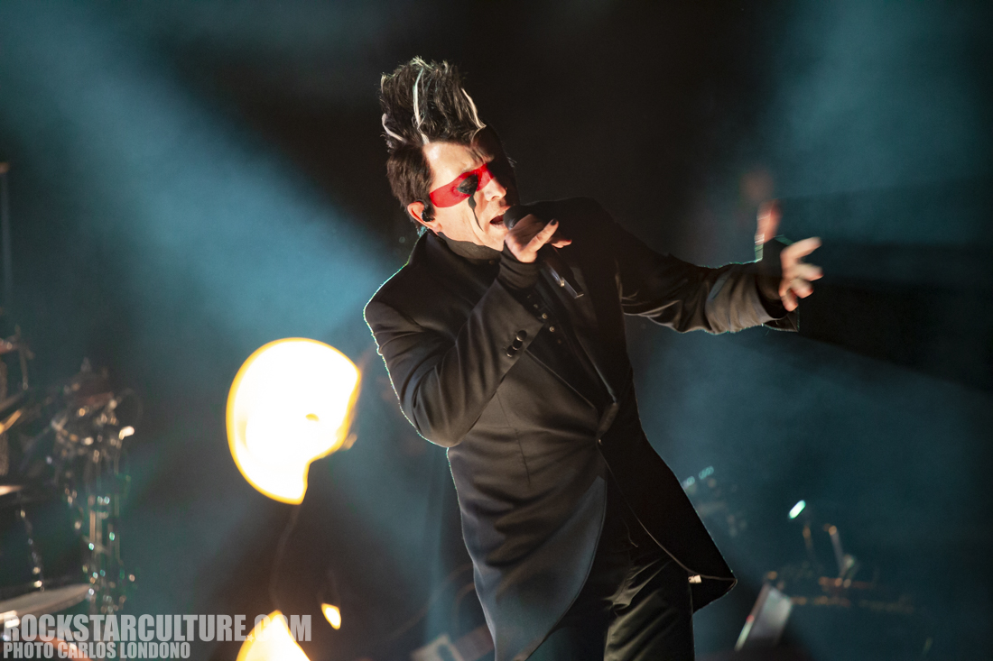 A male singer performing on stage with dramatic lighting, wearing a black suit and red sunglasses, passionately engaging with the audience.