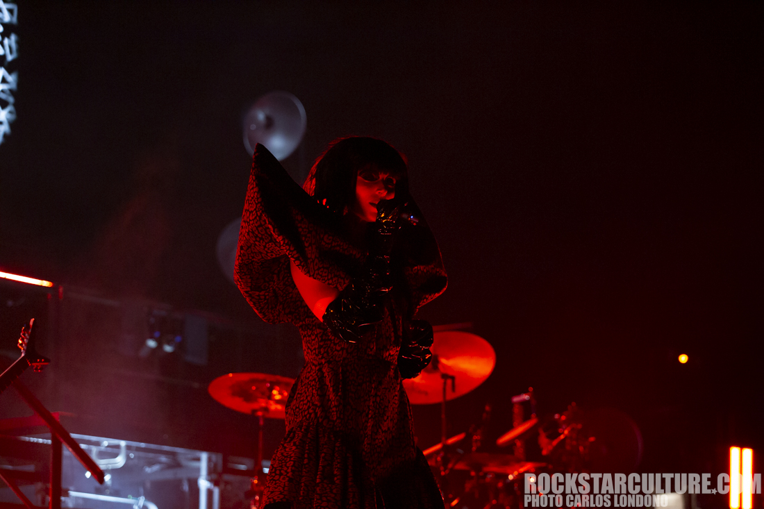 A performer in dark attire stands on stage with dramatic lighting, holding a microphone and captivating the audience.