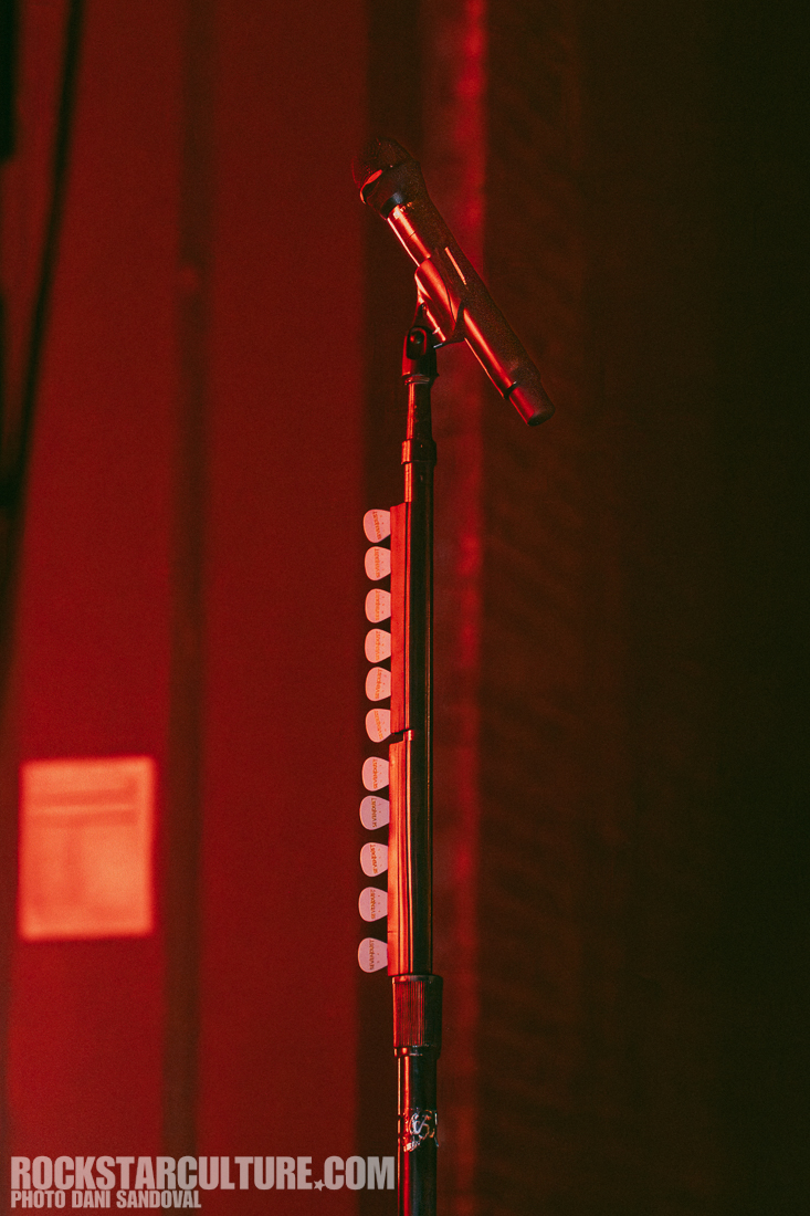 A microphone stand partially in shadow, with a black microphone on top and several guitar picks attached to the side, illuminated by red lighting.