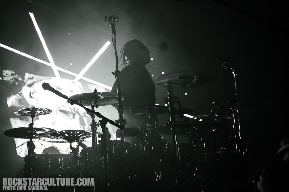 A silhouetted drummer performing on stage with drum set in a concert setting, illuminated by dramatic lighting and a backdrop of abstract visuals.