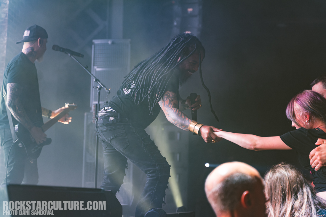 A performer with dreadlocks interacts with a female audience member during a live concert, reaching out to her as other band members play in the background.