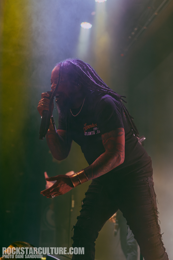 A male performer singing on stage, wearing a black t-shirt and holding a microphone. He has long dreadlocks and is dressed in stylish attire, surrounded by vibrant stage lights and a smoky atmosphere.
