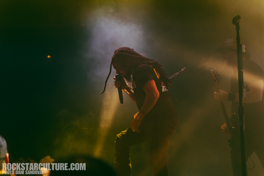 A musician with long dreadlocks performs on stage, holding a microphone and singing, with dramatic lighting and smoke effects in the background. Another band member can be seen partially behind them.