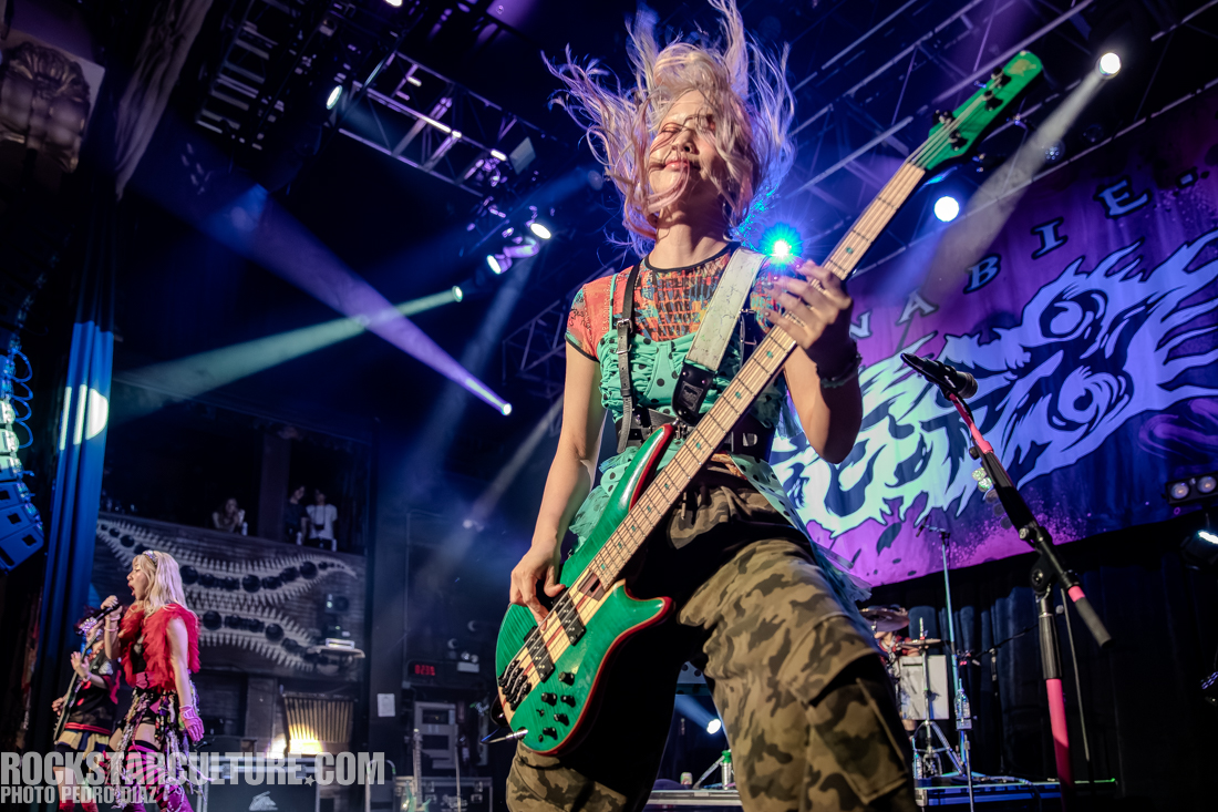 A female bassist performing on stage, energetically playing a green bass guitar with colorful stage lights in the background. The scene captures the excitement of a live music performance.