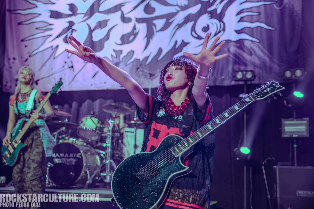 A lively performance by a musician holding an electric guitar and engaging with the audience, while another band member plays the bass in the background. The stage is adorned with a vibrant backdrop and lighting effects.
