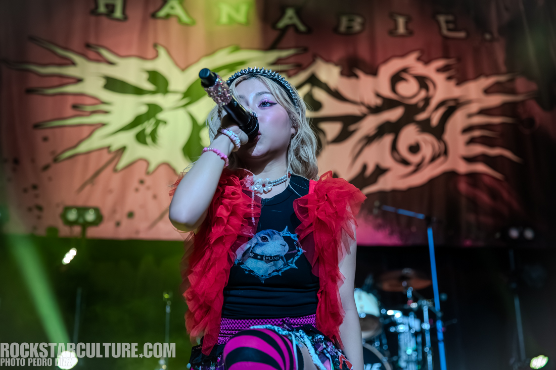 A young performer singing into a microphone on stage, wearing a black shirt with a graphic print, a red ruffled jacket, and colorful accessories, with a lively backdrop featuring an abstract design.