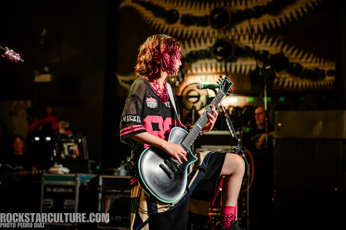 A musician playing an electric guitar on stage, wearing a black sports jersey and loose shorts, with pink socks and a vibrant hairstyle.
