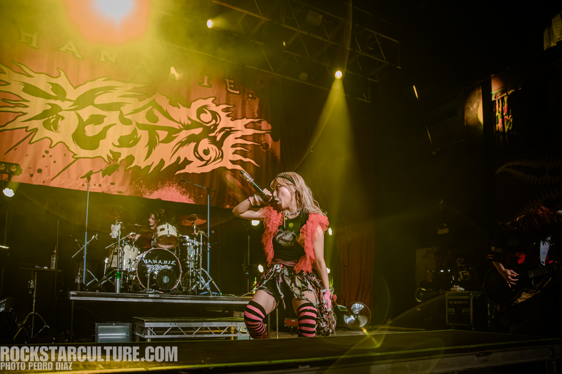 A lively rock concert scene featuring a female vocalist in a colorful outfit singing into a microphone, with a drummer visible in the background. The stage is illuminated with dramatic lighting and a large banner is displayed behind the performers.