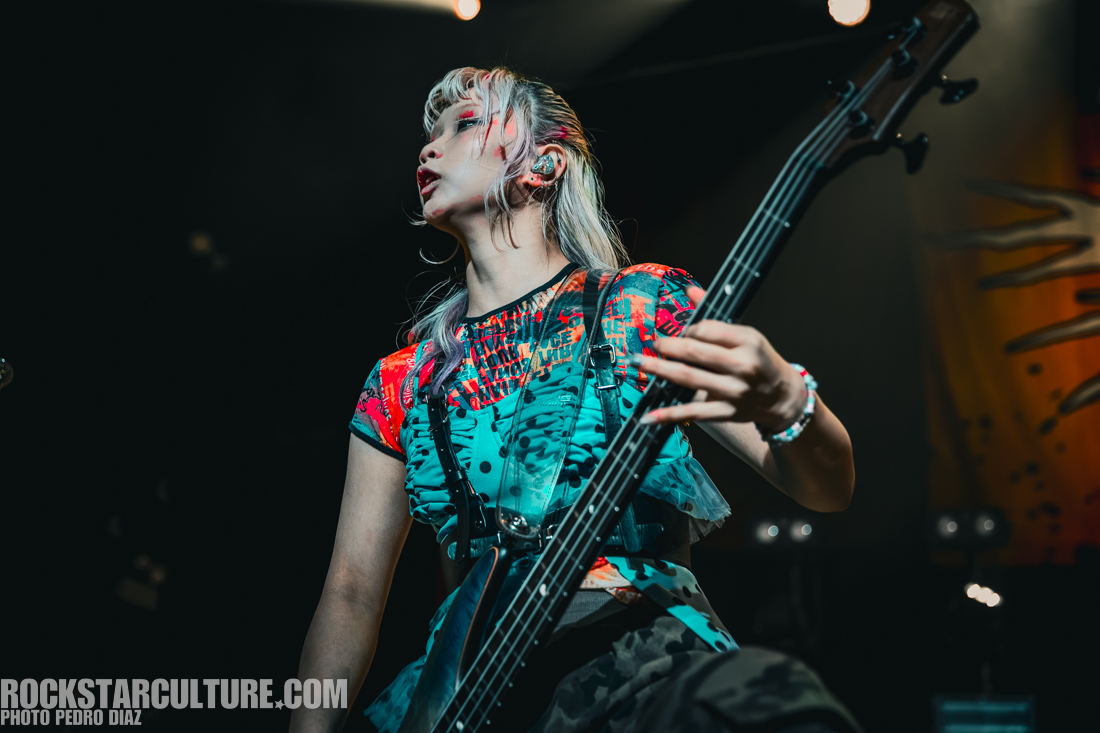 A musician playing a bass guitar on stage, wearing a colorful outfit with vibrant patterns, and performing with a focused expression.