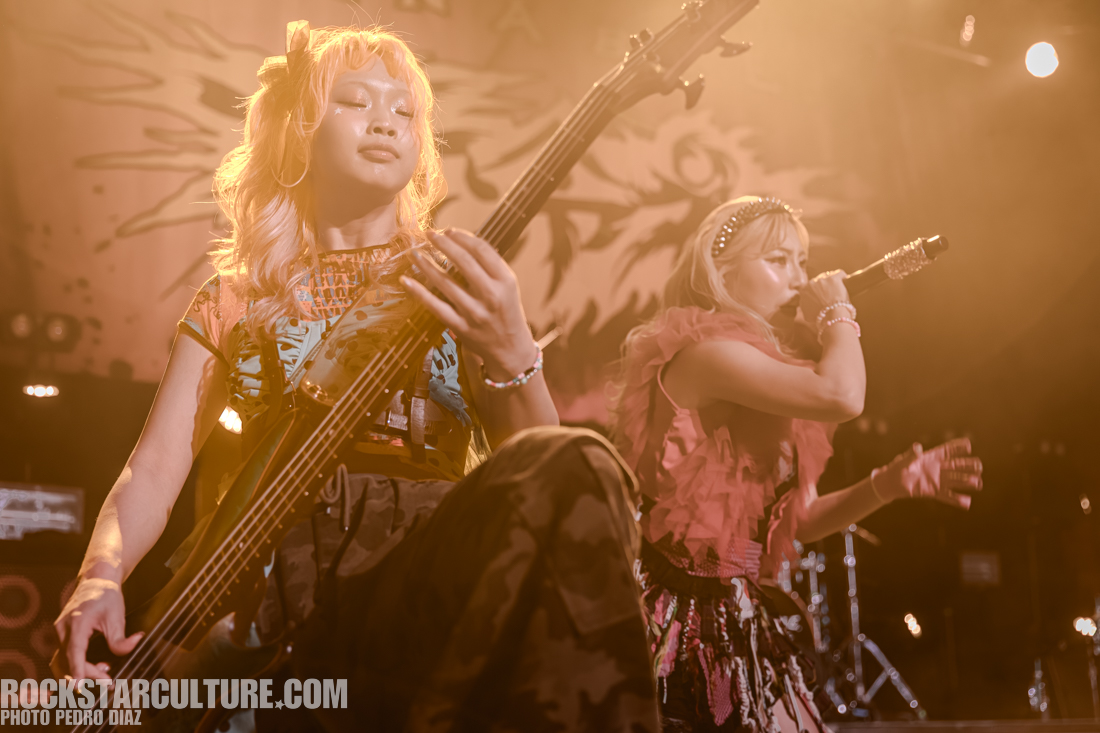 Two female musicians performing on stage; one is playing a bass guitar while the other sings into a microphone, surrounded by dramatic lighting.