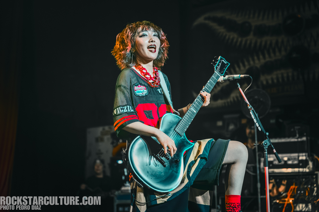 A musician performing on stage, playing an electric guitar and wearing a colorful oversized jersey and shorts, with a joyful expression.