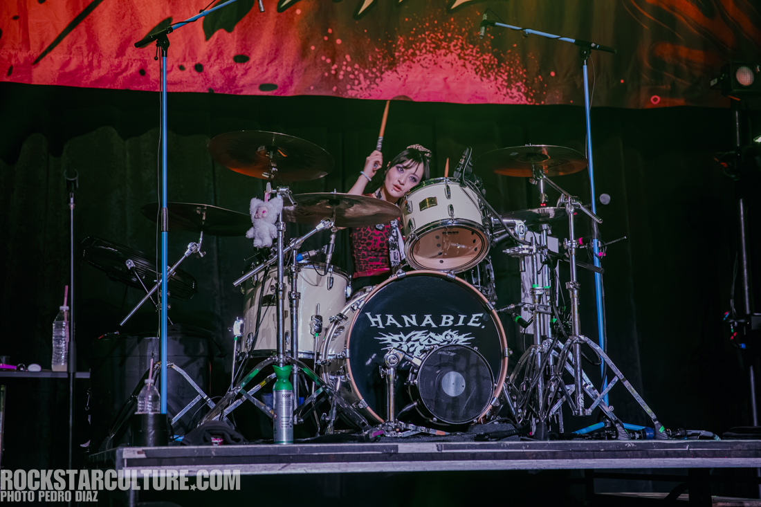 A drummer performs on stage with a vibrant backdrop, featuring a large 'HANABIE' logo on the bass drum. The drummer, wearing a black and red outfit, smiles while holding a drumstick.