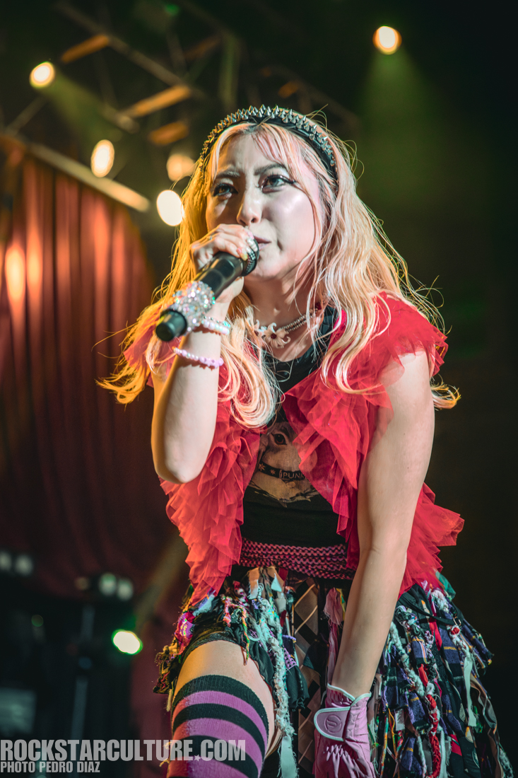 A singer on stage wearing a vibrant red ruffled top, black shirt, and colorful skirt, holding a microphone and performing passionately under stage lights.