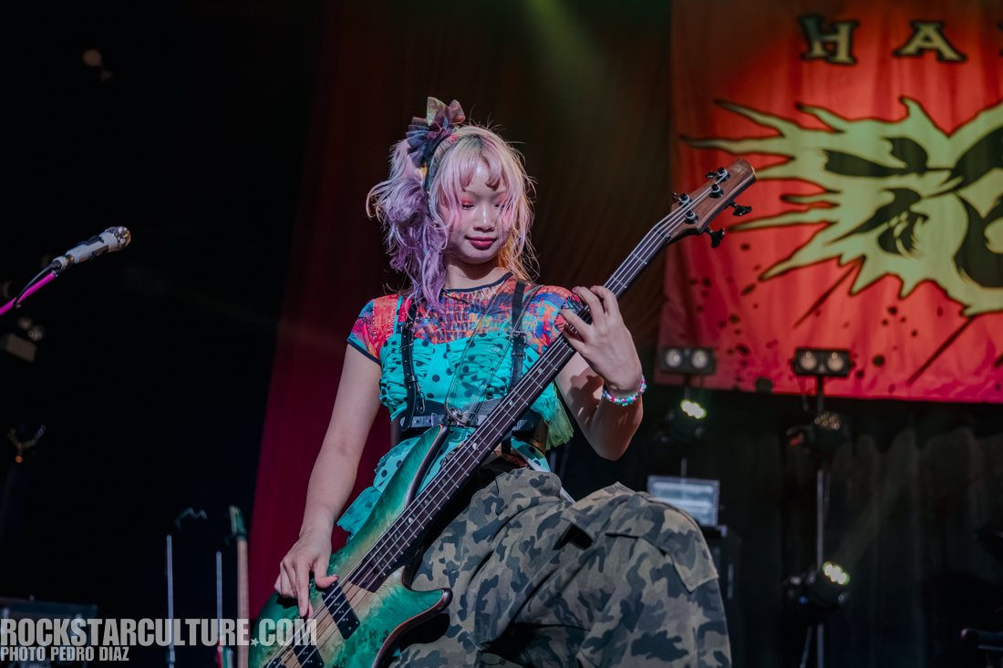 A musician playing a green bass guitar on stage, wearing a colorful outfit and camo pants, with vibrant purple hair and a bow accessory.