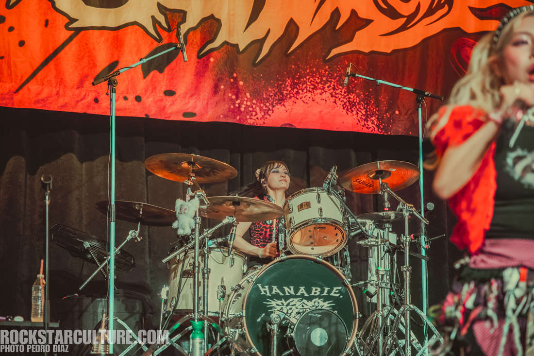 A drummer performing on stage with a colorful backdrop, featuring a drum set labeled 'HANABIE' and playful decorations.