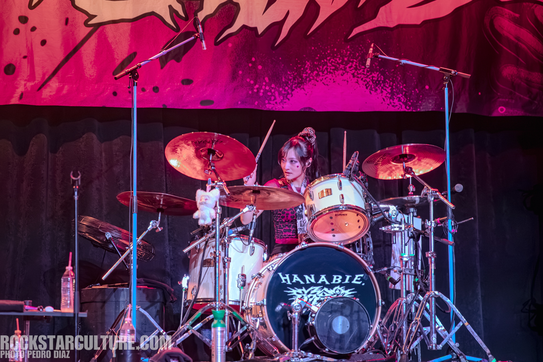 A female drummer performing on stage with a drum set, featuring the brand 'HANABIE'. She is surrounded by colorful lighting and a backdrop.