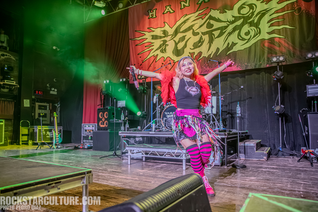 A performer on stage, dressed in a vibrant outfit with a pink top and colorful accessories, entertaining the crowd under green stage lights.