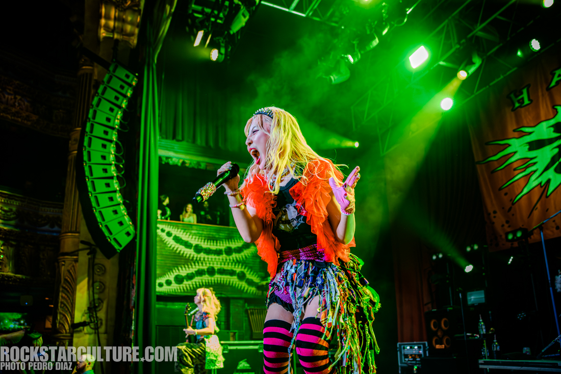A performer with long blonde hair and colorful attire sings passionately into a microphone on stage, surrounded by green lights. The stage features a vibrant backdrop and a second performer can be seen in the background.