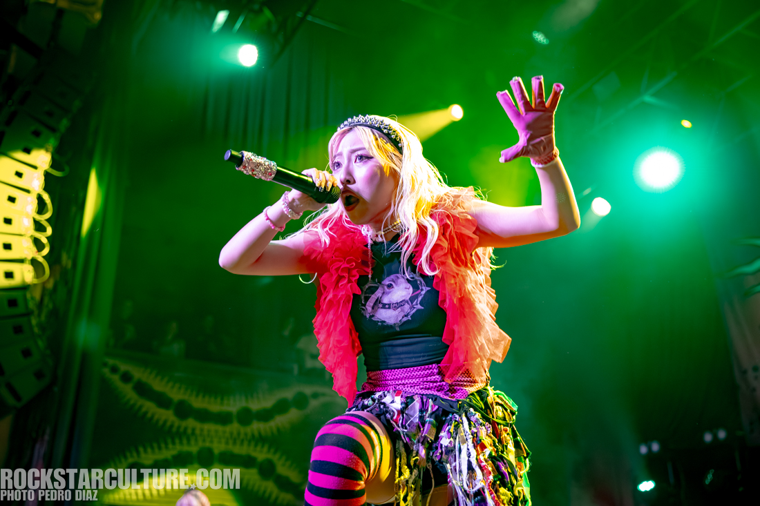 A female performer singing passionately on stage, wearing a vibrant outfit with a pink ruffled top and striped leggings, surrounded by colorful stage lights.