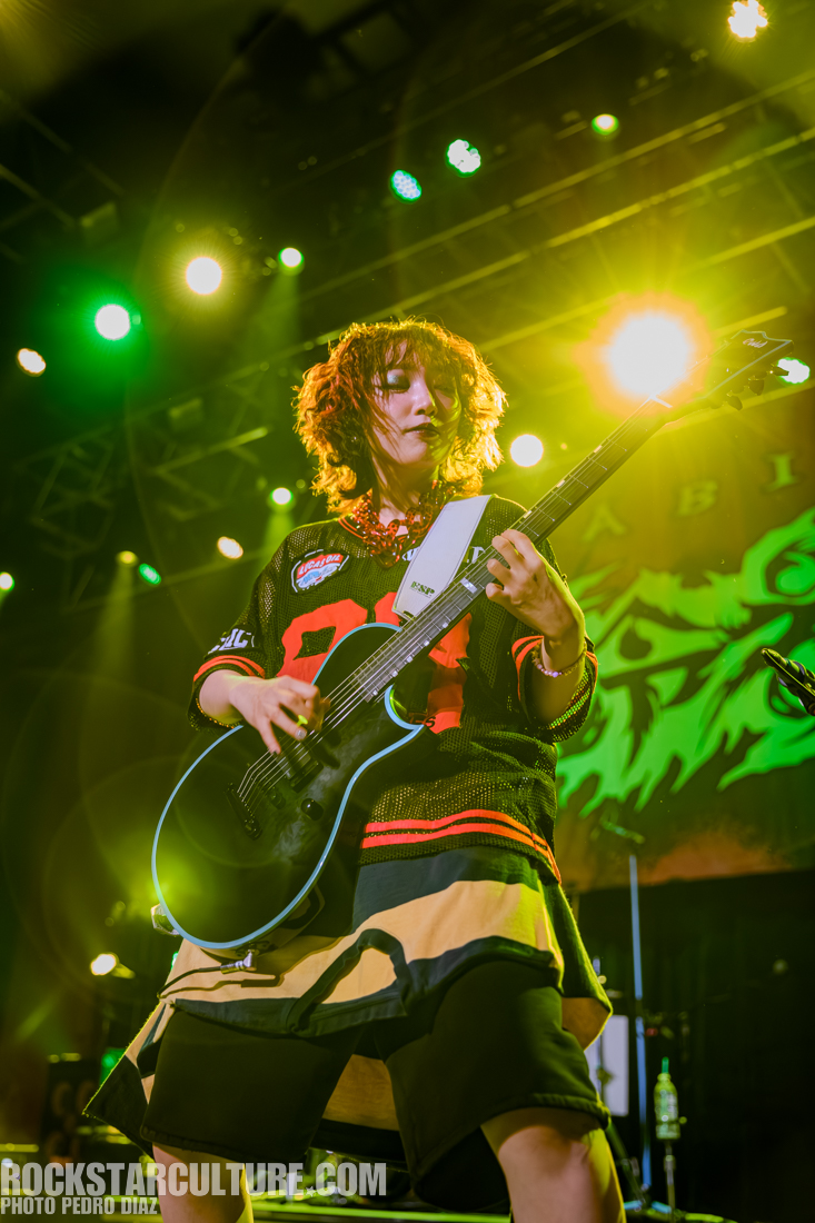 A musician performing on stage, playing an electric guitar, wearing a colorful oversized jersey and a distinctive skirt, with vibrant stage lights in the background.