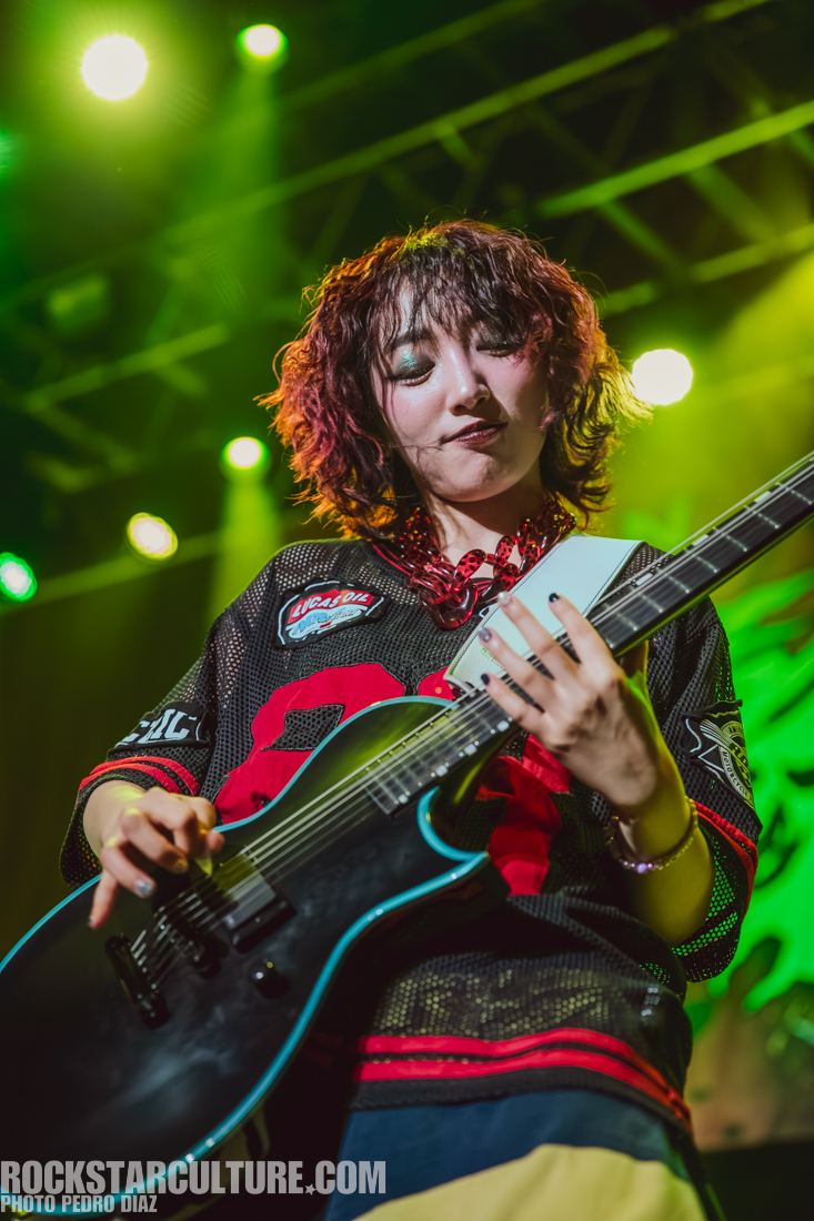 A musician with curly, dyed hair playing an electric guitar on stage under bright stage lights, wearing a black jersey with red accents.