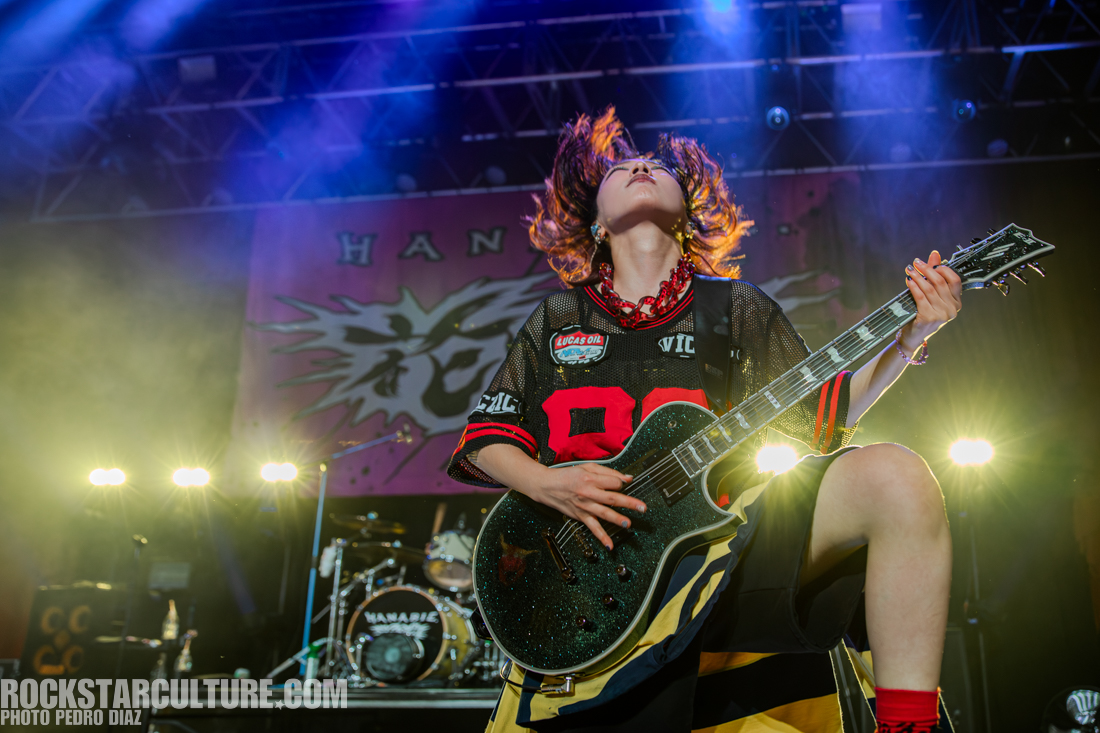 A musician passionately playing an electric guitar on stage, with their hair flying as they perform, wearing a basketball jersey and colorful accessories, illuminated by stage lights with a vibrant background.