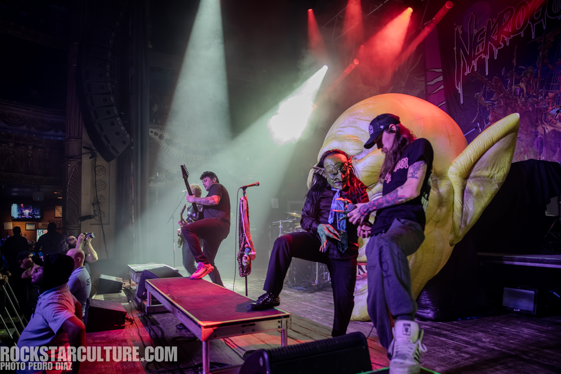 A live concert scene featuring three musicians performing on stage, with one wearing a green goblin costume. The background includes dramatic lighting and a large, cartoonish face sculpture.